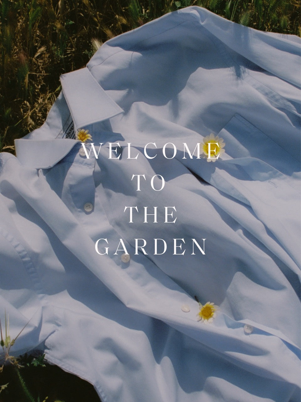 white shirt lying on grass with daisies on it. Copy reads Welcome to the garden.